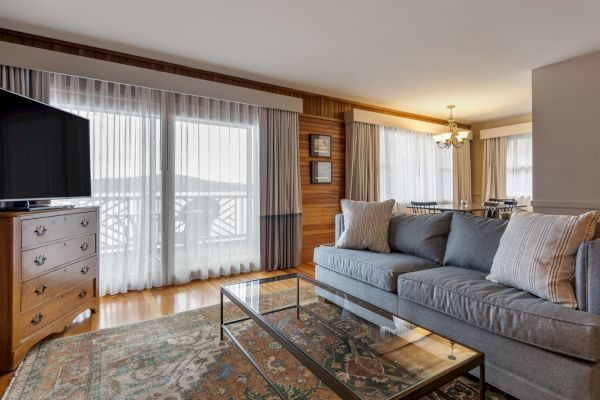 A cozy living room with a gray couch, glass coffee table, flat-screen TV, and large windows with sheer curtains, connected to a dining area.