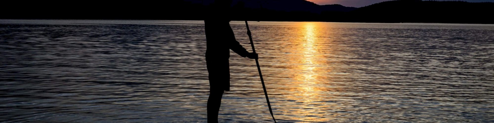 A person is paddleboarding on a calm lake during sunset, with mountains silhouetted in the background. The sky shows a mix of colors from the setting sun.