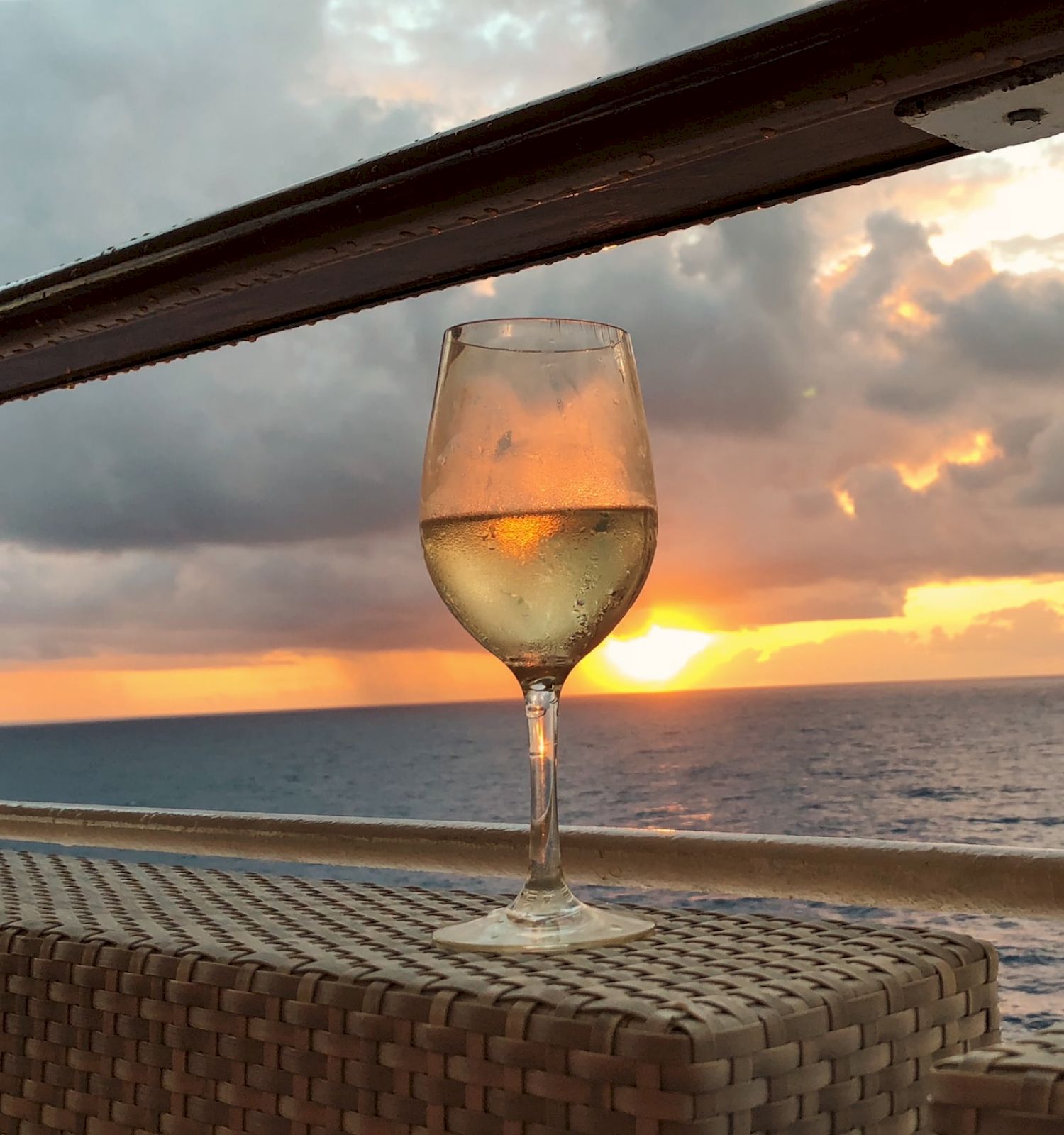 A glass of white wine sits on a wicker surface, with an ocean sunset in the background viewed from a balcony.