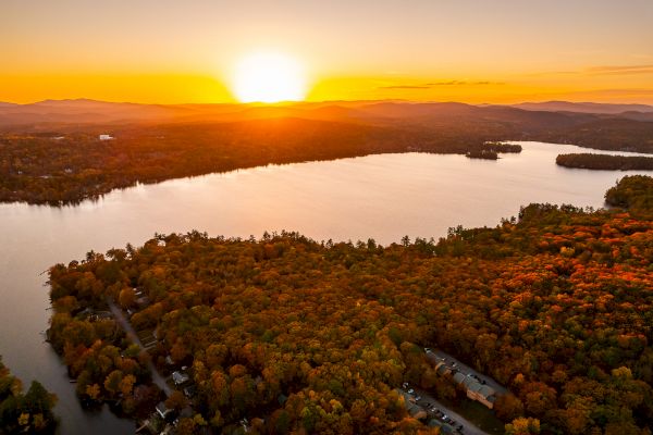 Aerial view of a serene lake surrounded by autumn foliage under a vibrant sunset, with distant mountains on the horizon.