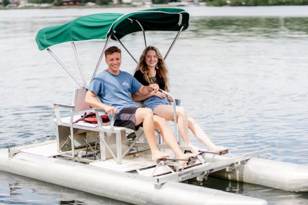 Two people are smiling while sitting on a pedal boat with a green canopy, floating on a calm body of water.