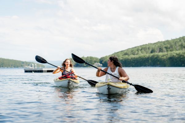 Two people are kayaking on a calm lake, surrounded by green hills and a clear sky. They seem to be enjoying a sunny day outdoors.