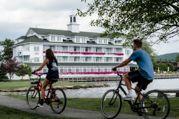 Two people are riding bicycles on a path by a lake with a large white building in the background, adorned with pink flowers.