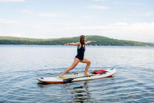 A person is practicing yoga on a paddleboard in a lake, balancing in a warrior pose with scenic hills and a clear sky in the background.