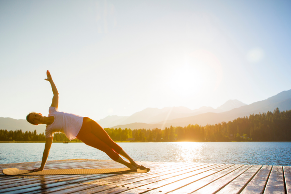 A person performs a yoga pose on a dock by a lake, with mountains and a setting sun in the background, under a clear blue sky.