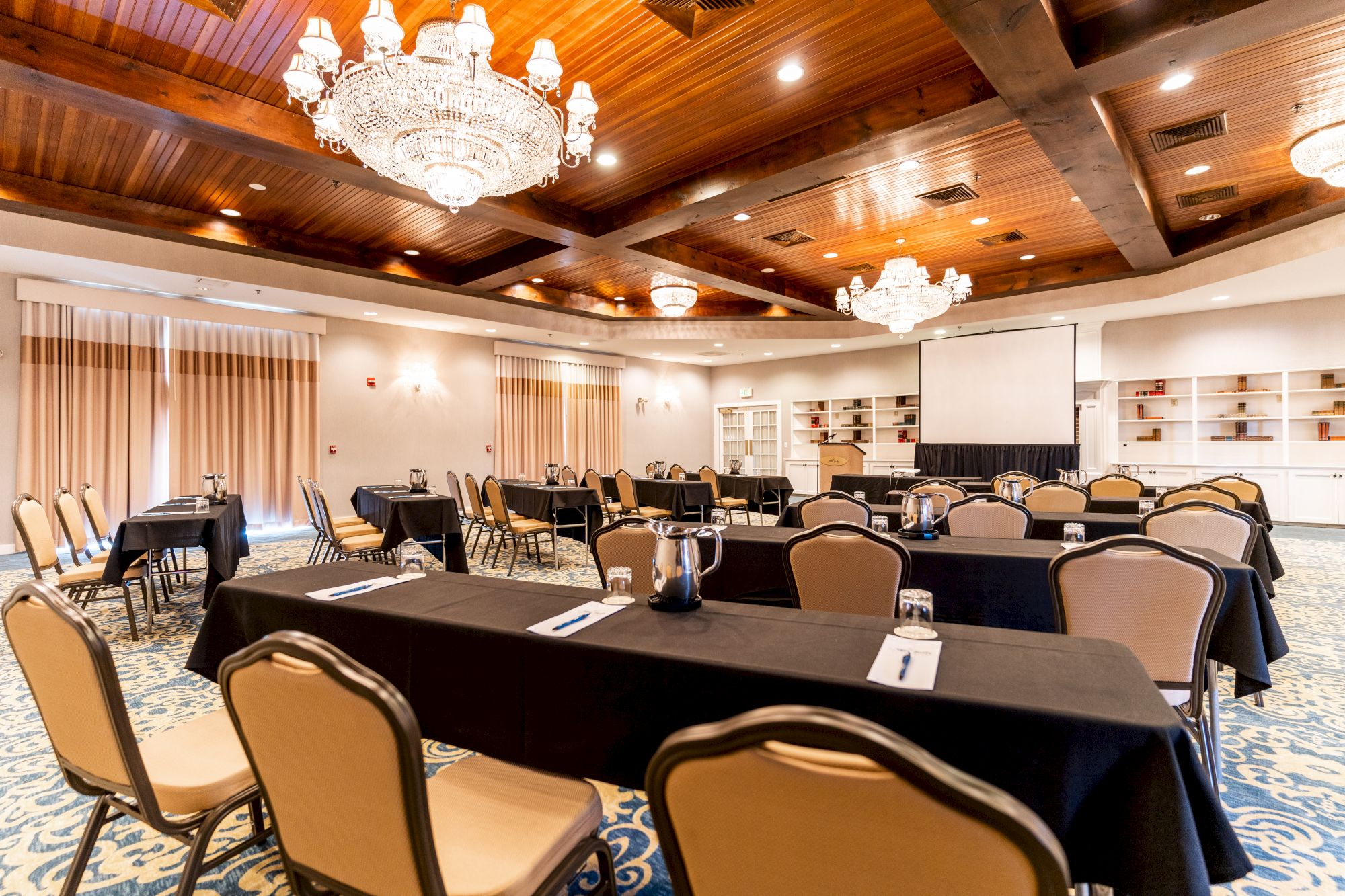 The image shows a well-lit conference room with chandeliers, arranged tables, chairs, and a large screen for presentations.