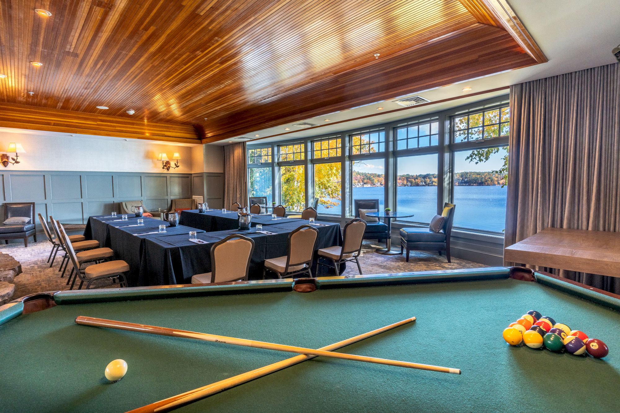 A room with a pool table in the foreground, conference tables, chairs, and a scenic window view of a lake and trees in the background.