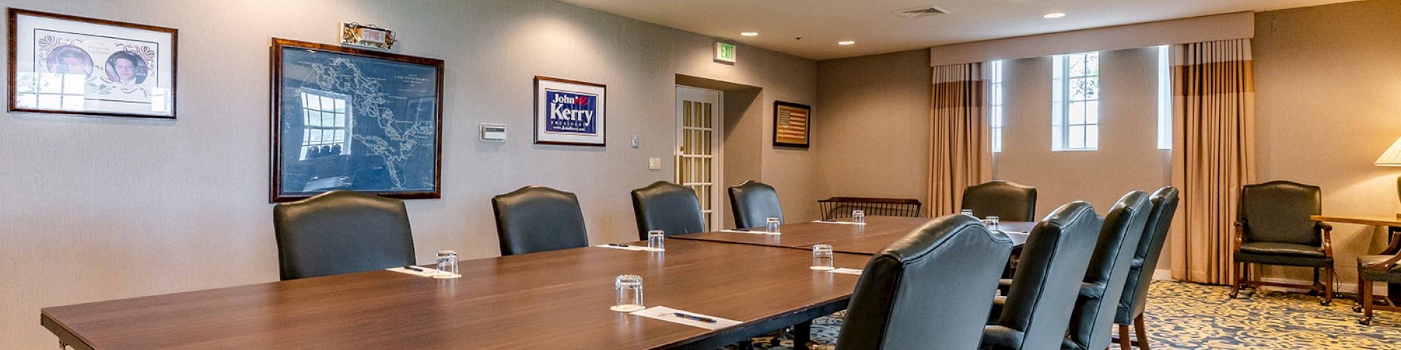 A conference room with a large table, padded chairs, framed artwork, patterned carpet, and windows with curtains.