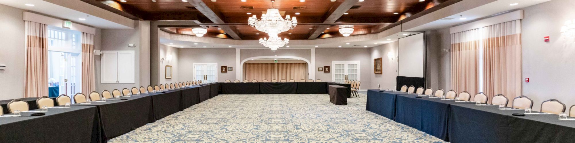 A spacious, elegant conference room with long tables arranged in a U-shape, chandeliers, and a patterned carpet, ready for a meeting or event.