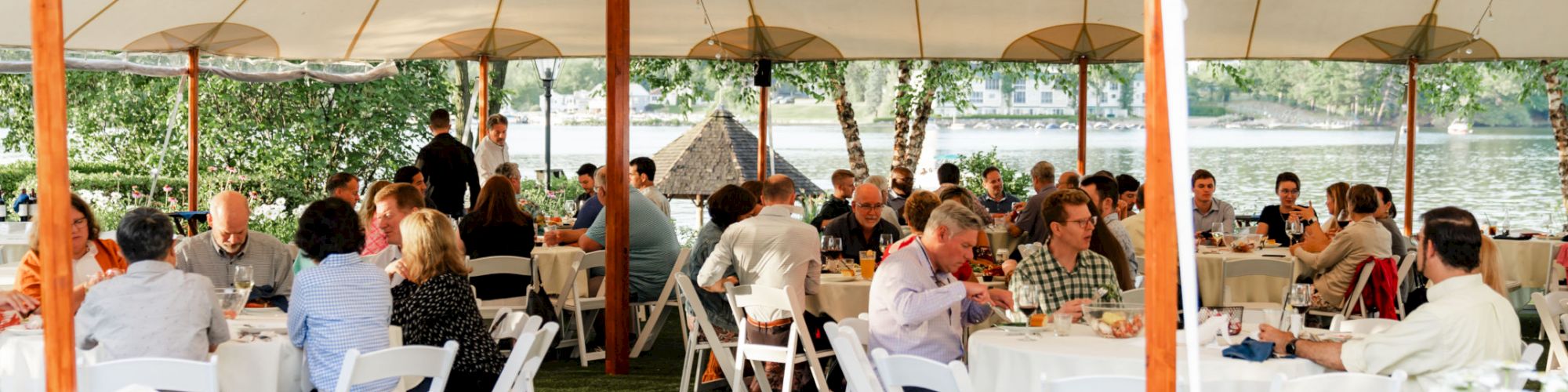 People are seated at round tables under a large outdoor tent, engaged in conversation. The setting appears to be a social or formal event.