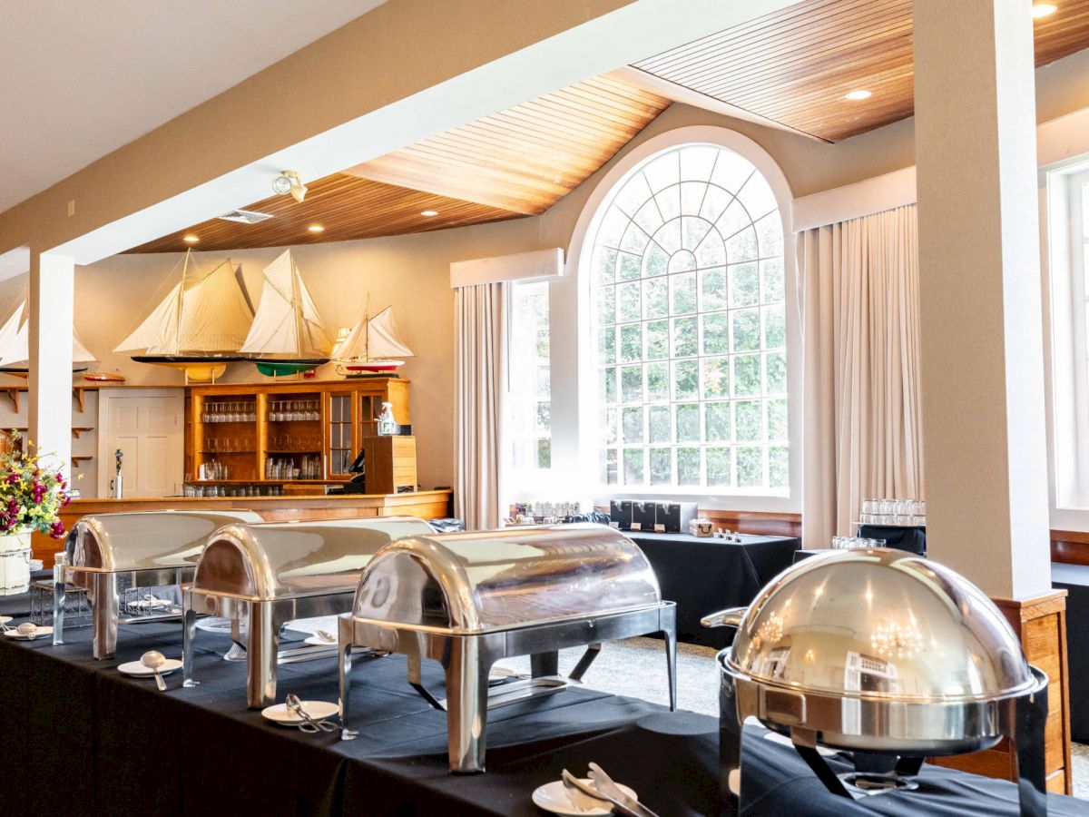 A buffet setup in a room with large arched windows, model sailboats on a shelf, and chafing dishes on black tablecloths.