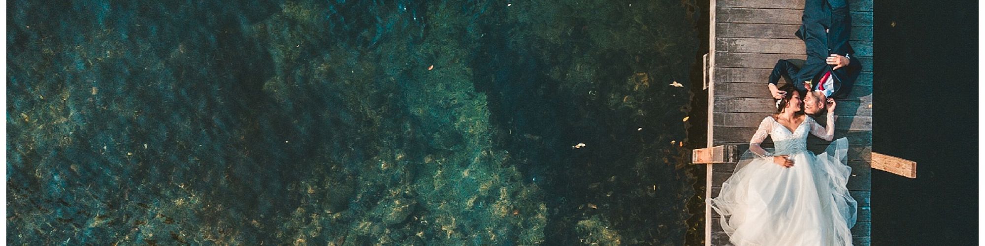 A couple in wedding attire is lying on a dock above clear water, sharing a romantic moment.