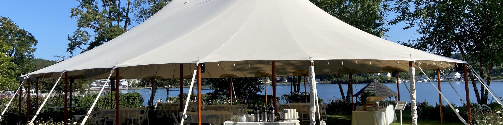 A large canopy tent is set up outdoors on a grassy area near a lake, with tables and chairs underneath and surrounded by trees and flowers.