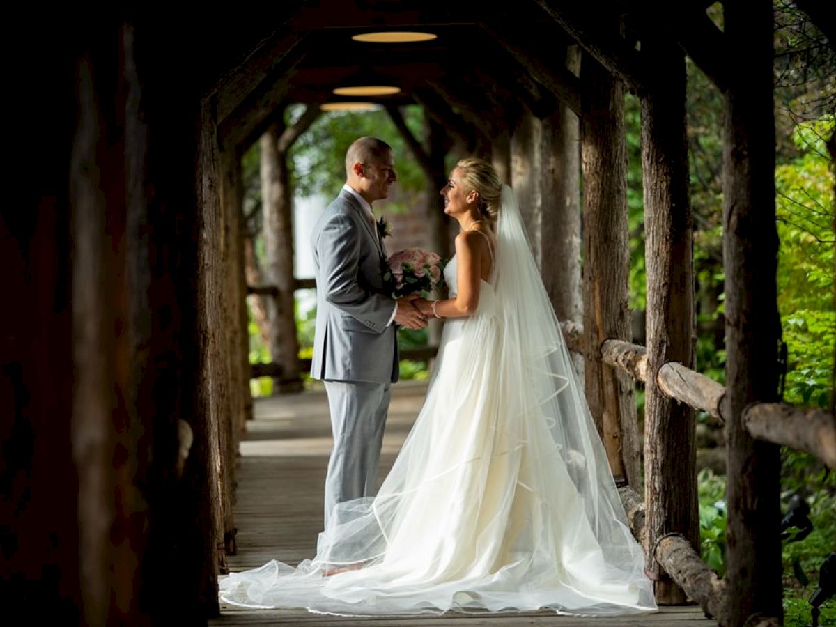 A couple is standing on a wooden bridge, dressed in wedding attire, facing each other while holding hands and smiling.