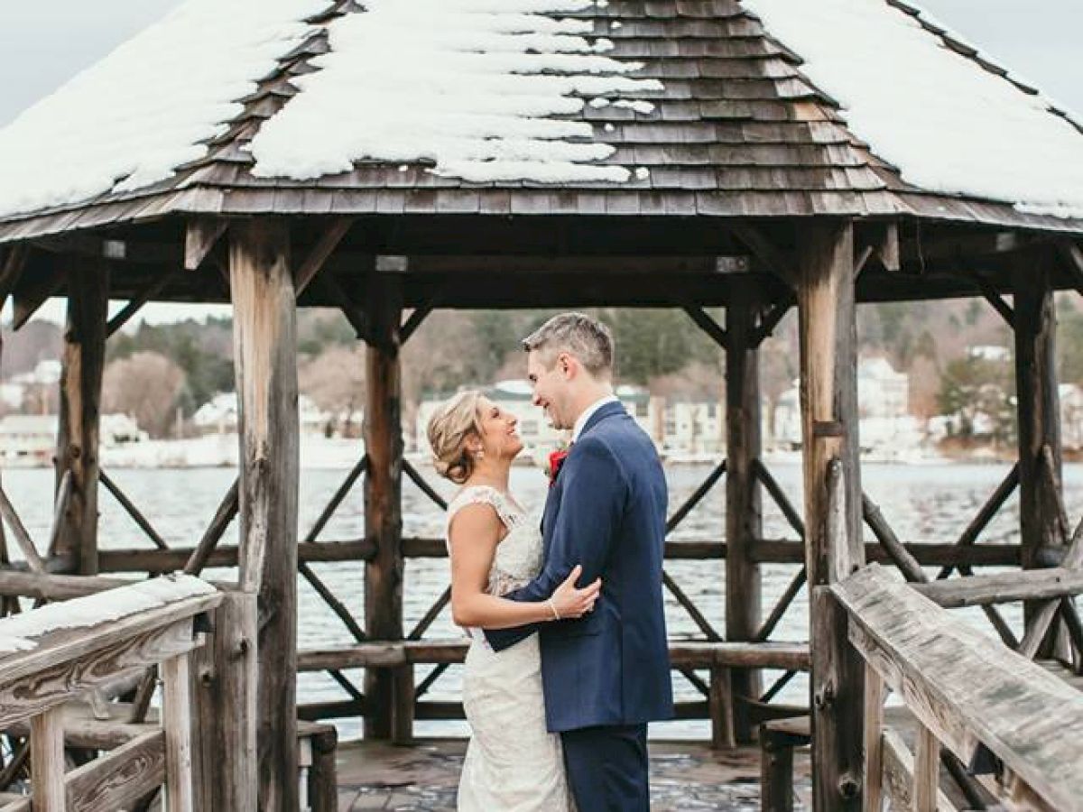 A couple in wedding attire embraces under a snowy gazebo on a pier. The groom wears a blue suit, and the bride is in a white dress.