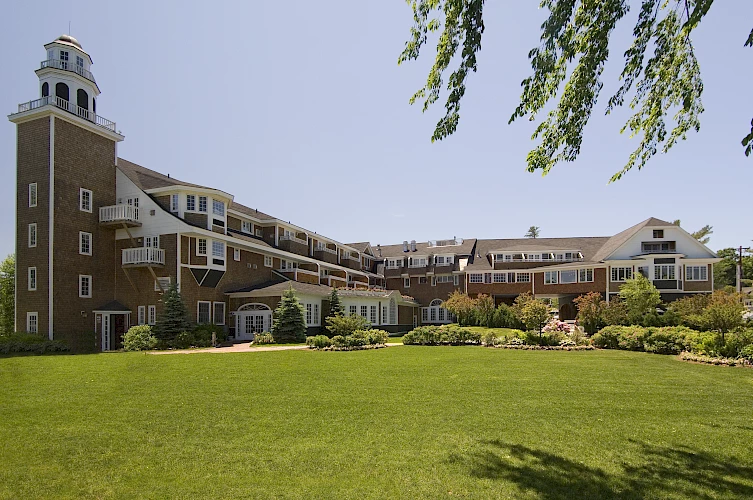 A large, multi-story building with extensive landscaping and a tower on one side is shown, set against a clear blue sky and a green lawn.