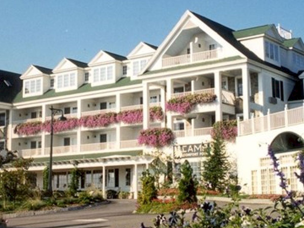 The image shows a large, multi-story building with balconies adorned with pink flower baskets, surrounded by greenery and featuring a well-kept garden.