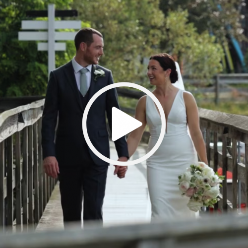 A couple in wedding attire is walking hand in hand on a wooden bridge, smiling at each other. The bride holds a bouquet of flowers.