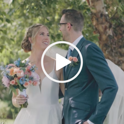 A bride and groom are smiling at each other outdoors. The bride holds a bouquet of flowers, and the groom is in a suit.