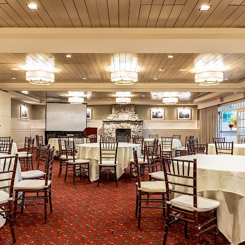 A spacious banquet hall with round tables, white tablecloths, wooden chairs, a projector screen, and a stone fireplace under a wooden ceiling.