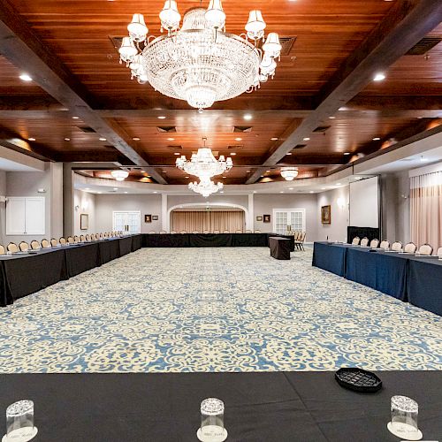 A large, elegant conference room with chandeliers, U-shaped table setup, ornate carpet, and black tablecloths, prepared for a meeting or event.