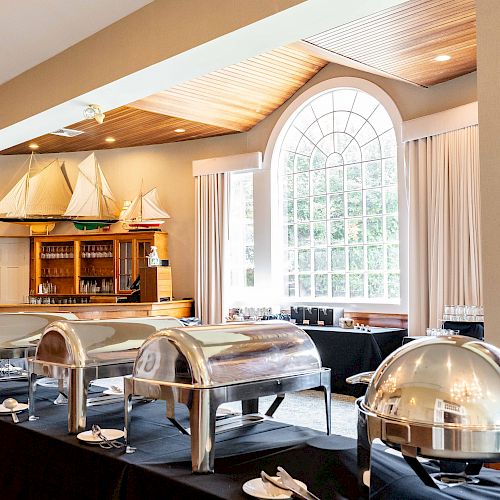 A well-lit dining area features a buffet setup with chafing dishes and a large window in the background, and sailboat models on display.