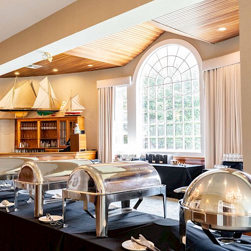 A buffet setup with stainless steel chafing dishes and a flower arrangement, in a room with large windows and model sailboats on display.