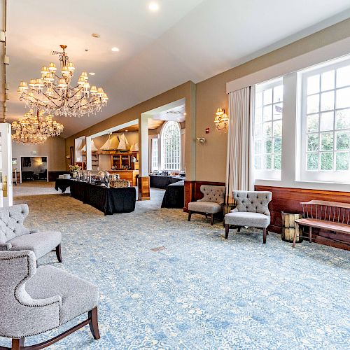 The image shows a spacious, elegant room with chandeliers, large windows, upholstered chairs, a wooden bench, and a buffet setup in the background.