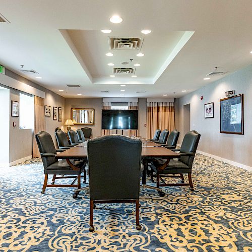 A well-lit conference room with a long table, surrounded by chairs, decorative carpet, framed pictures on the walls, and a large TV screen in the back.