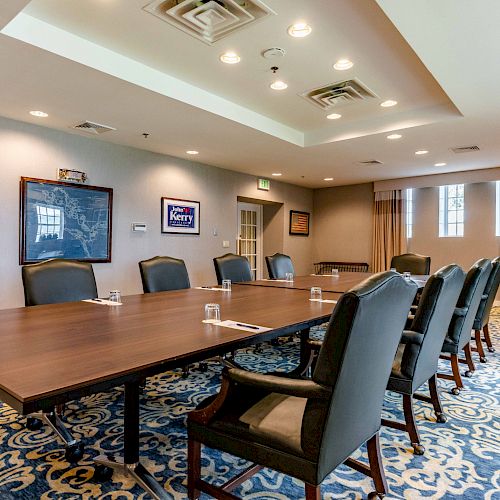 A conference room with a long table, chairs, framed pictures on the wall, and patterned carpet. The room is well-lit with ceiling lights.