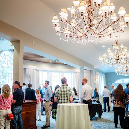 People socializing in an elegant room with chandeliers, tables, and a bar area, seemingly at a social event or gathering.