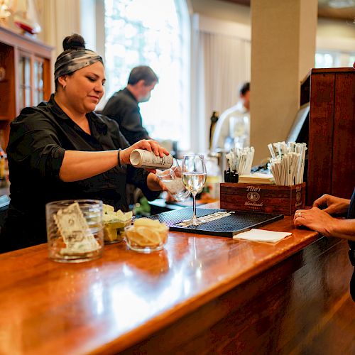 A bartender is pouring a drink for a customer at a wooden bar counter, with other patrons and staff in the background.