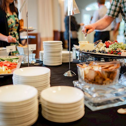 A buffet setup with plates, fruits, vegetables, and dips, with people serving themselves in the background.