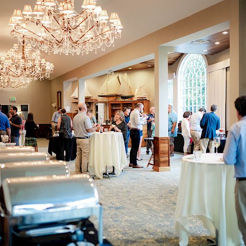 People are mingling at an indoor social event with chandeliers, high tables, and food stations, set under elegant lighting in a spacious room.