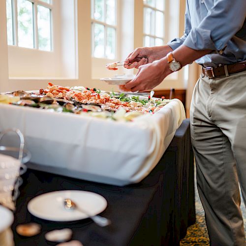 A person is serving food from a buffet table with a variety of dishes, including seafood and vegetables, in a well-lit room.