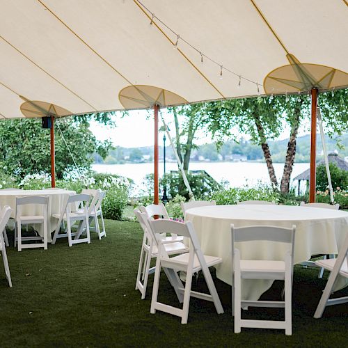 An outdoor event setup under a tent with round tables covered in white cloth and surrounded by chairs, overlooking a scenic waterfront view.