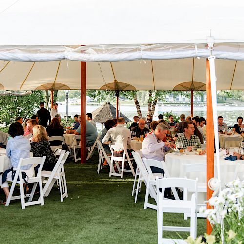 A group of people are gathered under a large outdoor tent, seated at round tables with white chairs, in a garden-like setting near a body of water.