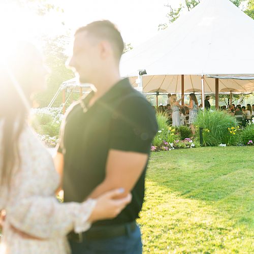 A couple standing in a garden, embracing. In the background, there is a large, white canopy tent with people gathered underneath.