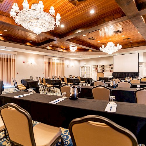 A conference room with chandeliers, tables covered with black cloths, and several chairs arranged in rows and clusters, ready for a meeting.