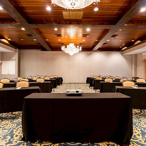 A conference room with rows of tables and chairs, a projector on a table, chandeliers, and patterned carpeting is shown.