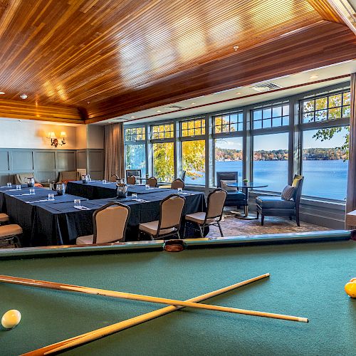 This image shows a room with a billiards table in the foreground and a meeting area with chairs and tables by large windows overlooking a lake.