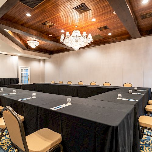 A U-shaped conference room setup with chairs and black table covers, decorated with chandeliers and wooden ceiling accents, ready for a meeting.