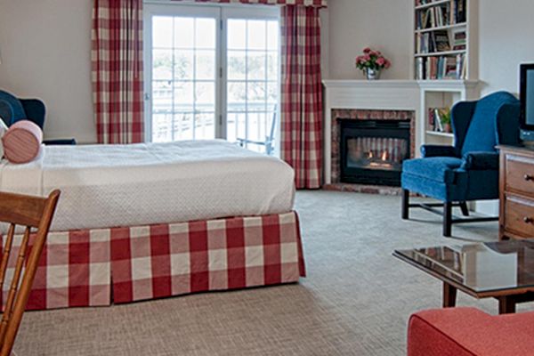 A cozy bedroom with a bed, desk, armchairs, and a fireplace, featuring a TV, bookshelf, and red-checkered curtains near a window.