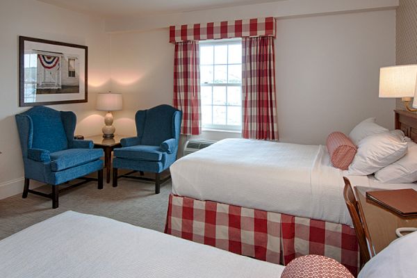 The image shows a cozy hotel room with two beds, checkered skirts, and pillows. There are blue chairs, a window with red-checkered curtains, and two lamps.