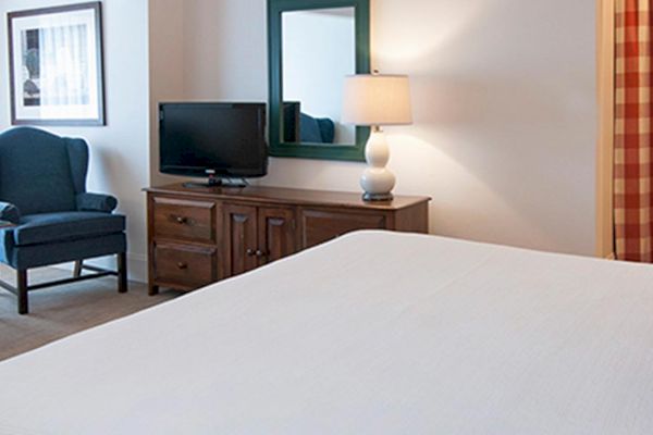 A neat, cozy hotel room with a bed, two armchairs, lamps, TV, and a checkered curtain. There's a table between the chairs and a dresser under the TV.