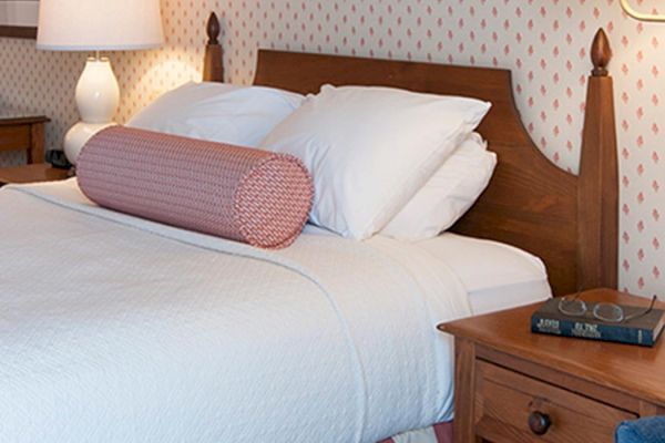 A neatly made bed with white bedding and a pink bolster pillow beside a wooden nightstand with a book and glasses, and a blue chair to the right.