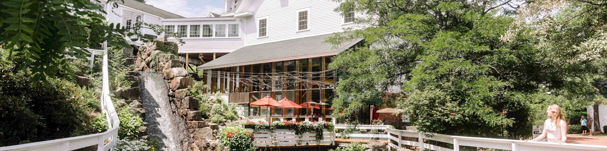 The image shows a charming white building with a cascading waterfall and outdoor seating beside a stream, surrounded by lush greenery and trees.