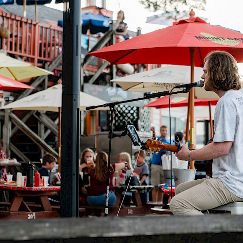 A musician is performing outdoors under red umbrellas, with people watching from picnic tables in a vibrant, busy setting, ending the sentence.