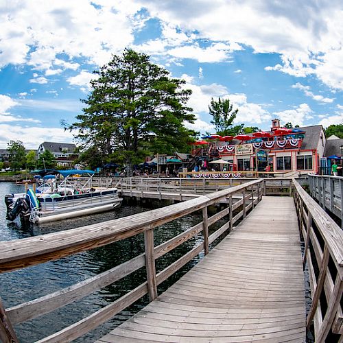 A scenic waterfront with a wooden boardwalk, boats docked, and a lively building adorned with festive decorations under a partly cloudy sky.