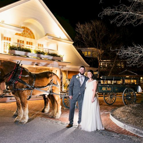 A couple in formal attire stand in front of a horse-drawn carriage outside a brightly lit building at night.